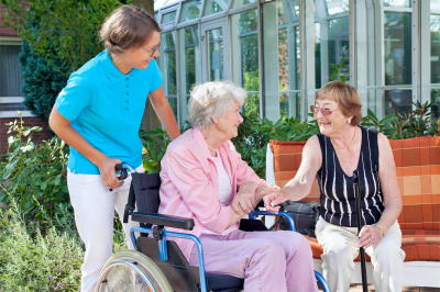 Elderly woman on a wheelchair with her caregiver talking to a friend who is sitting on a wooden garden bench